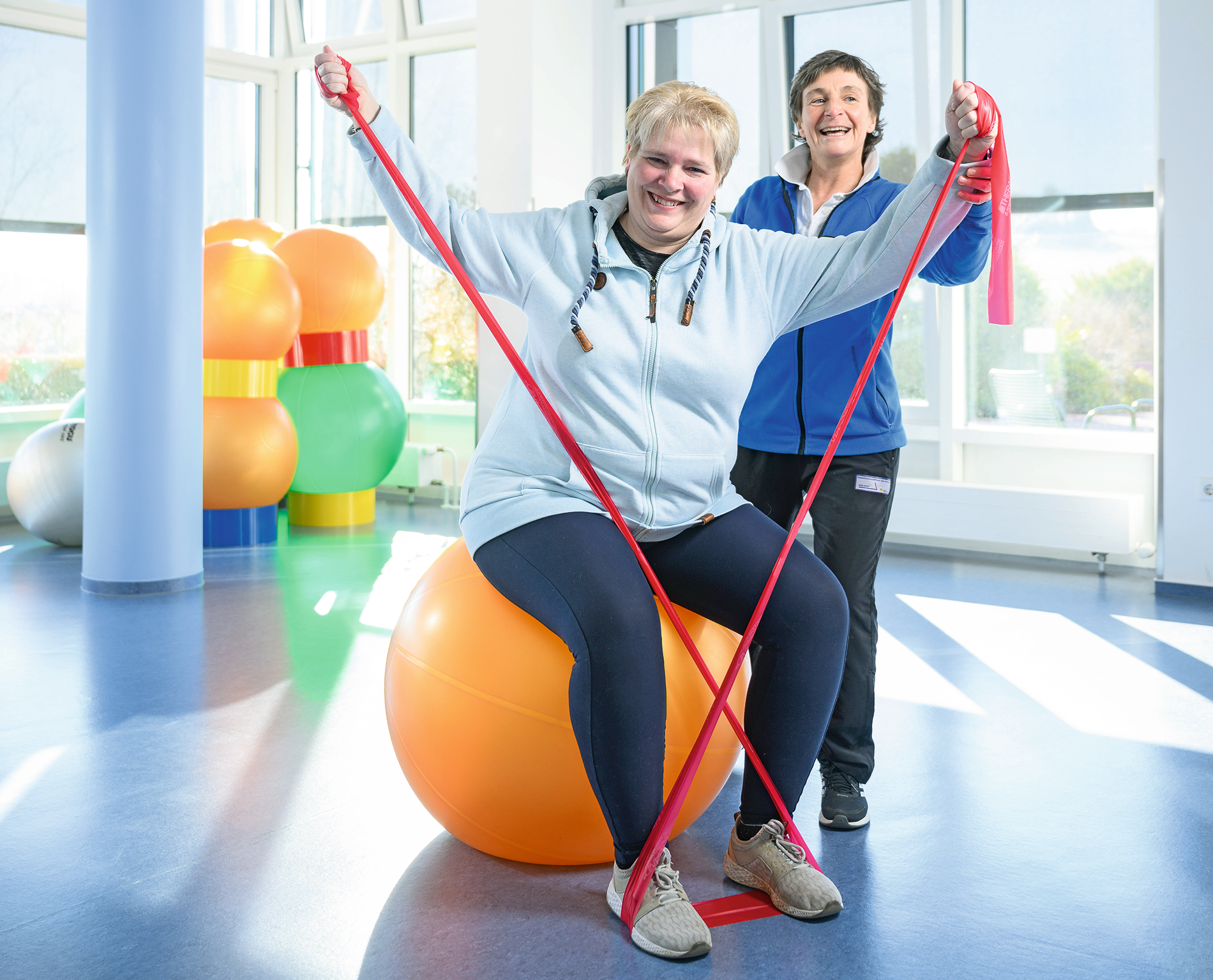 Nina Seegers sitzt lachend auf einem Gymnastikball und macht Übungen mit einem Theraband. Eine Physiotherapeutin leitet sie an.