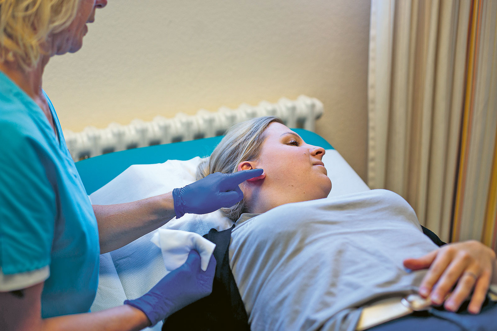 Behandlerin mit Handschuhen deutet auf das Ohr einer auf einer Liege liegenden Patientin und hält ein Tuch bereit.