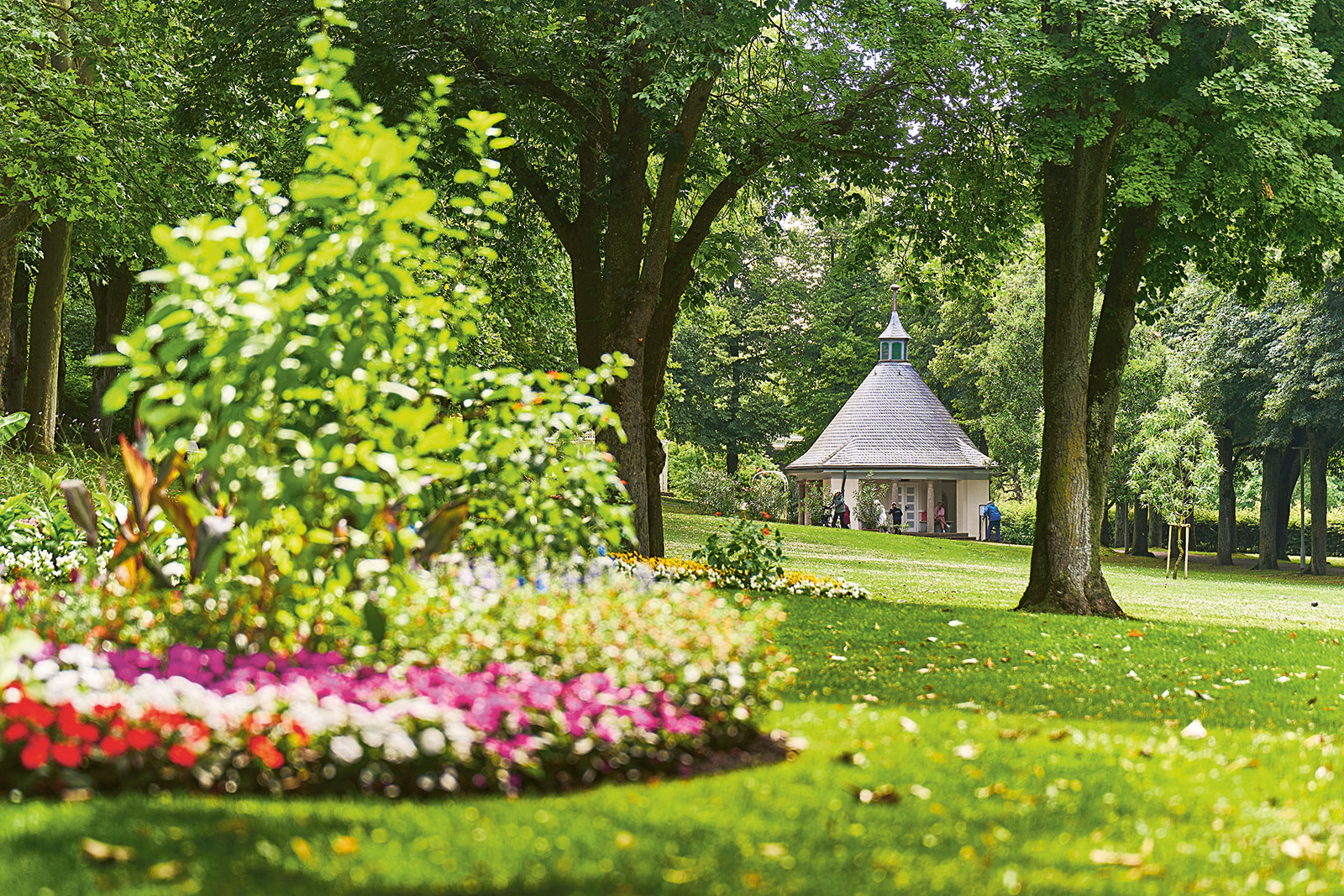 Grüne Parklandschaft mit großen Bäumen und bunten Blumenbeeten im Vordergrund; in der Mitte steht ein kleiner weißer Pavillon mit spitzem Dach, daneben sind einige Menschen zu sehen.