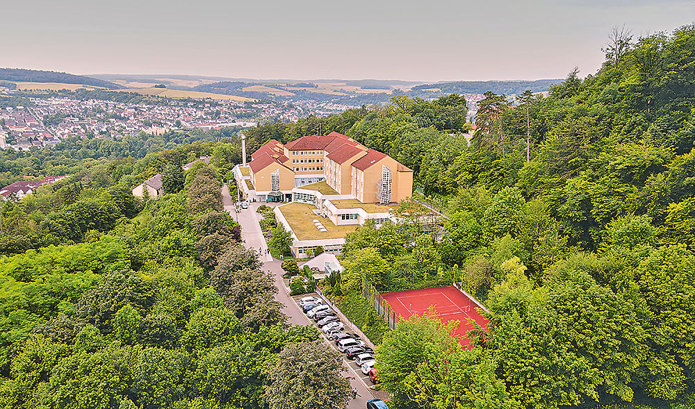 Luftaufnahme eines großen Gebäudekomplexes mit roten Dächern am bewaldeten Hang; unten ein Parkplatz und ein roter Tennisplatz, im Hintergrund Blick auf eine Stadt und Felder.
