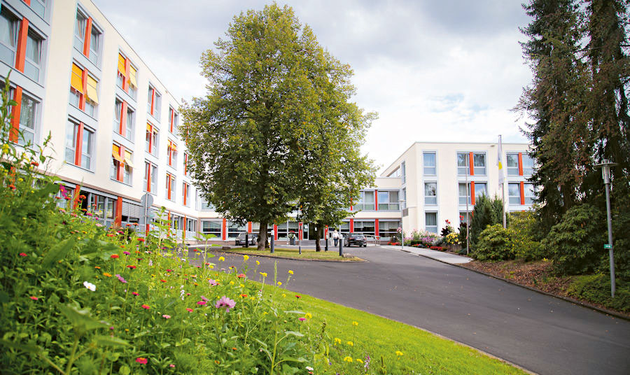 Außenansicht eines modernen Gebäudekomplexes mit mehreren weißen Gebäuden und orangefarbenen Fensterakzenten; im Vordergrund eine Blumenwiese und eine asphaltierte Zufahrt, in der Mitte ein großer Baum.